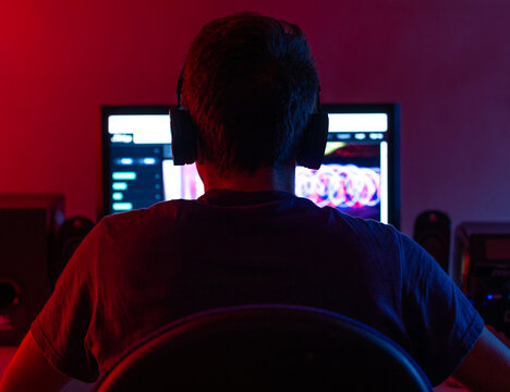 Back view of calm young brunet in casual wear and wireless headset browsing contemporary computer with blank screen while sitting at table in dark room
