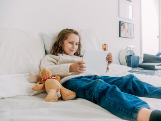 Smiling glad little girl wearing casual clothes lying on comfortable bed and watching video on modern tablet in light bedroom