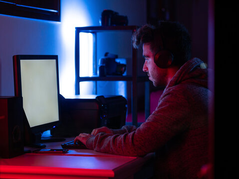 Side view calm young brunet in casual wear and wireless headset browsing contemporary computer with blank screen while sitting at table in dark room