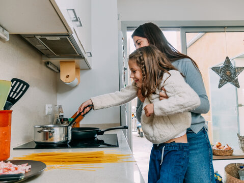 Cheerful young mother lifting cute smiling daughter stirring food on frying pan while cooking together in contemporary kitchen