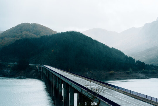 Picturesque scenery of road bridge over blue calm river flowing through forested hills on foggy day