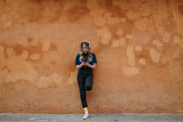 Young focused female in wireless headset text messaging on cellphone while standing with raised leg near wall in town on brown background