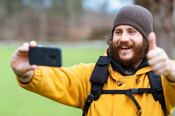 Happy adult bearded male traveler with thumb up taking self portrait on cellphone in daylight