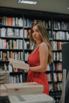 Side view of young female in red sundress with open textbook standing in bookshop and looking at camera