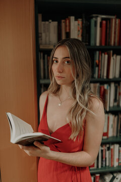 Side view of young female in red sundress with open textbook standing in bookshop and looking at camera