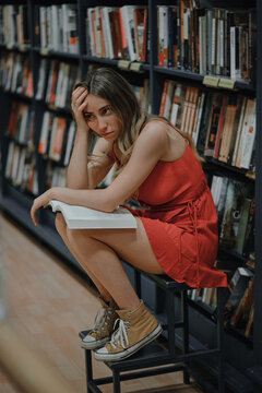 Side view of young exhausted female in sundress and gumshoes touching forehead while sitting with textbook and looking away in bookshop