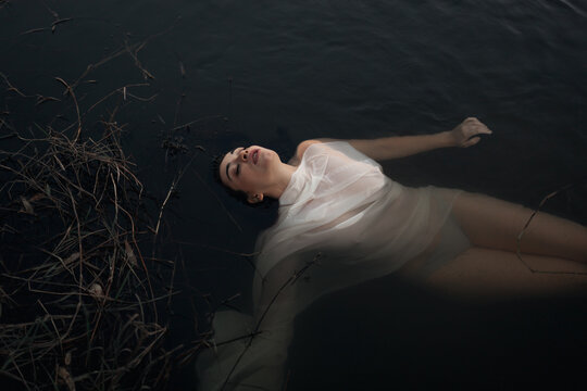 From above serene young brunette in wet panties and translucent white cloth lying with eyes closed on tranquil dark pond water