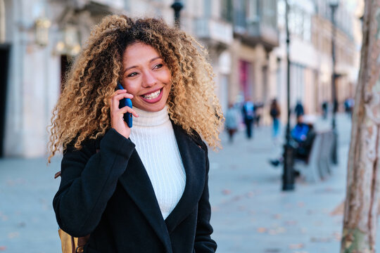 Happy young African American female with long curly hair smiling while chatting on mobile phone on urban street
