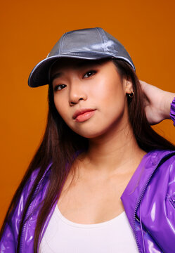 Portrait of Asian young woman with serious expression. She wears a purple jacket and a gray cap and is looking at the camera against a yellow background while touching her head.