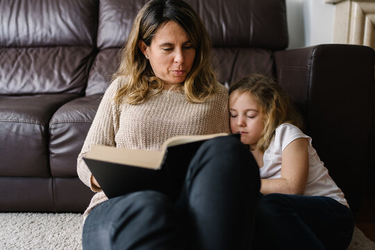 Adorable little girl cuddling mom and sleeping while mother reading book in living room at home