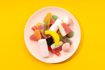 Top view of yummy multicolored gummy candies on striped plate placed on yellow background