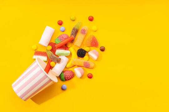 Top view of assorted multicolored sweet jelly candies and marshmallows scattered near disposable striped cup on yellow background