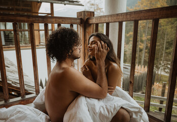 Romantic young undressed couple cuddling gently while sitting on wooden cottage porch and covering bodies with cozy white blanket on autumn weather