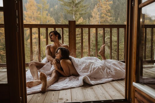 Romantic undressed couple lying on cozy blanket and cuddling on wooden cottage terrace against deciduous forest on autumn