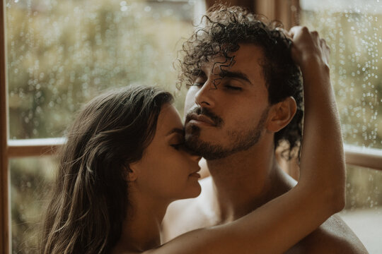 Tender woman embracing gently bearded boyfriend with closed eyes near window with water drops in shack