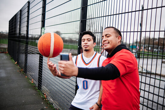 Latino friends taking a selfie by the fence of a basketball court outdoors. - Powered by Adobe