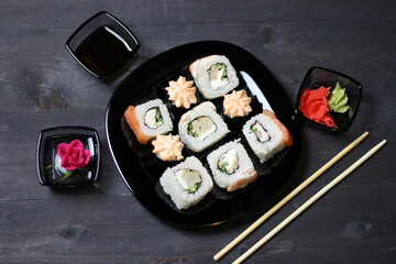 Pink rose in small bowl, marinated ginger and wasabi, soy sauce, rice rolls with salmon, crab, shrimp in plate and chopsticks on black background