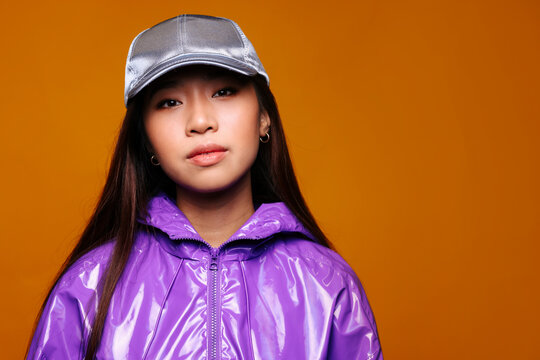 Portrait of Asian young woman with serious expression. She wears a purple jacket and a gray cap and is looking at the camera against a yellow background
