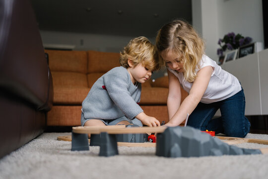 Cute little curly haired siblings sitting on floor and playing with toy road and cars while spending time together in living room at home