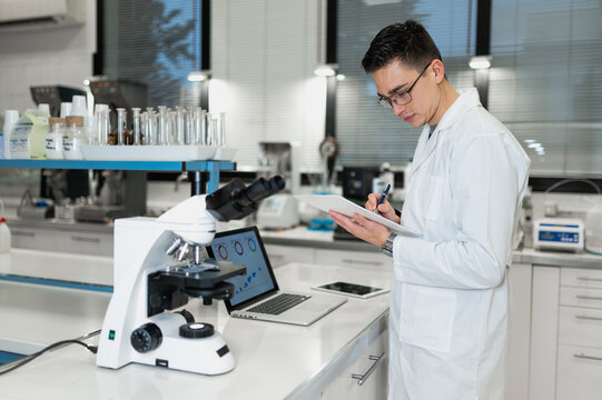Side view of young male microbiologist in white coat examining chemical sample through microscope and making notes in journal while working in modern laboratory