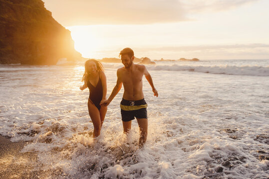 Full length of happy young couple in swimwear holding hands and walking together in waving sea water at sunset time while enjoying holidays on Tenerife island in Spain