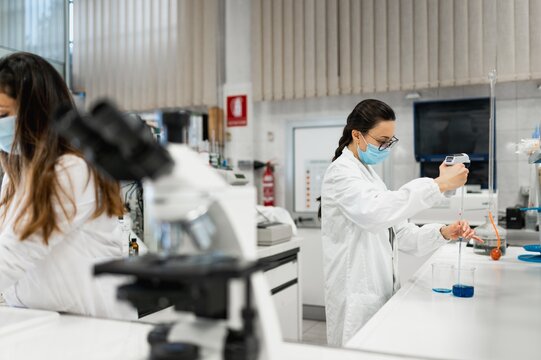 Professional female scientist in white coat and protective mask conducting chemical experiment with blue liquid in modern lab