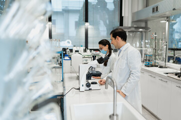 Happy male scientist using microscope and analyzing chemical samples while female colleague working on laptop in modern laboratory