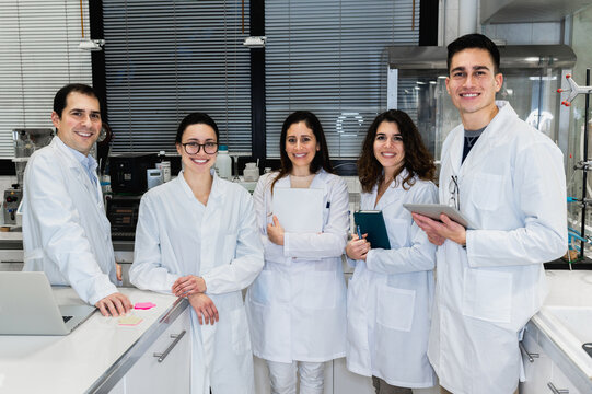 Group of positive young scientific researchers in white coats smiling and looking at camera while standing together in modern lab