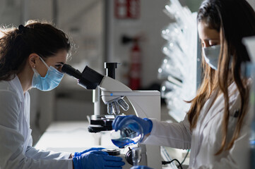 Young female biochemistry students in white coats and protective masks and gloves researching chemical reaction with microscope during experiment in modern lab