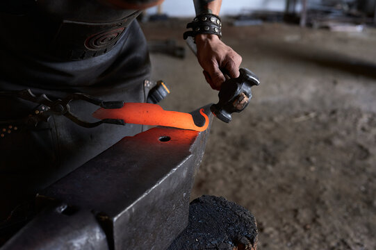 Side view of anonymous tattooed young male forger in apron heating metal in furnace while working in grungy smithy