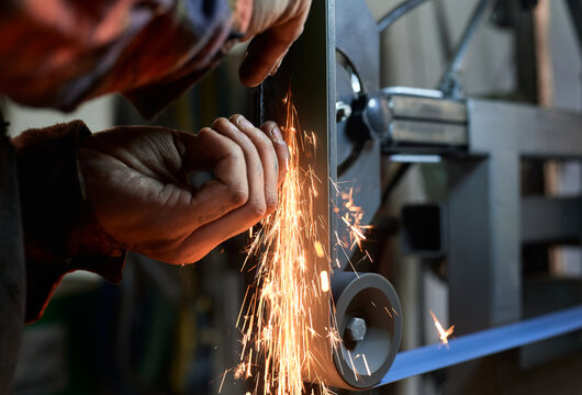 Side view of unrecognizable young tattooed male blacksmith in protective goggles and respirator polishing small metal details on grinder machine in workshop