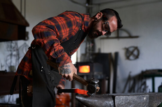 Side view of young brutal bearded craftsman in apron and goggles heating and striking iron on anvil with hammer during work in traditional smithy