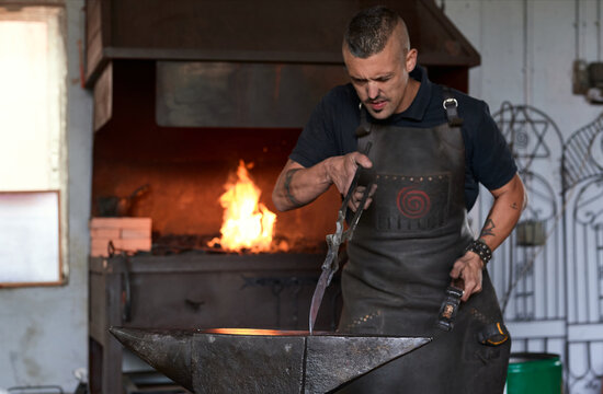 Concentrated young bearded blacksmith in apron and protective goggles striking heated metal with hammer on anvil while working in forge
