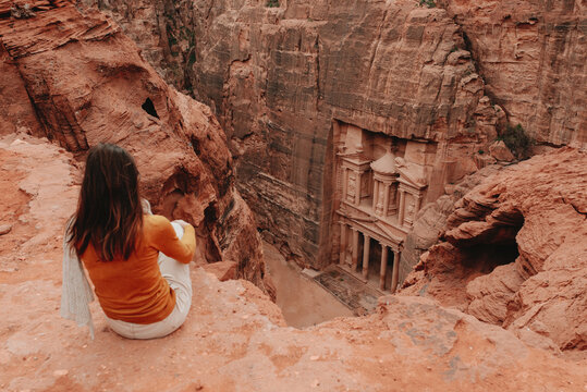 From above back view of anonymous female traveler in casual clothes enjoying view of historic Al Khazneh temple carved out of rock sitting on edge of cliff in Petra