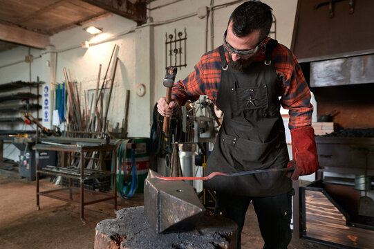Concentrated young bearded blacksmith in apron and protective goggles striking heated metal with hammer on anvil while working in forge
