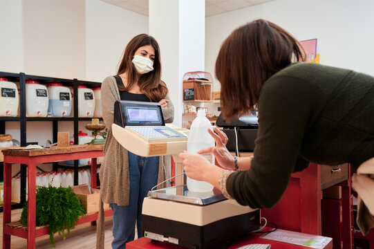 Side view of crop unrecognizable cashier weighing bottle of environmentally friendly bottle of detergent and pressing buttons on scales in shop for client standing at counter