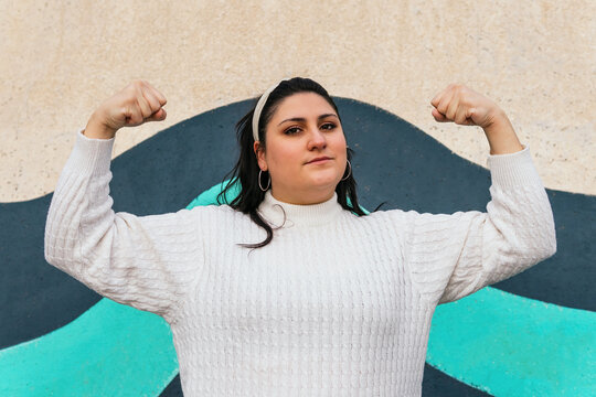 Young Powerful Plump Female In White Knitwear Standing With Raised Arms And Looking At Camera Outdoors