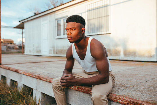 Young masculine African American male in undershirt with clasped hands looking away against house