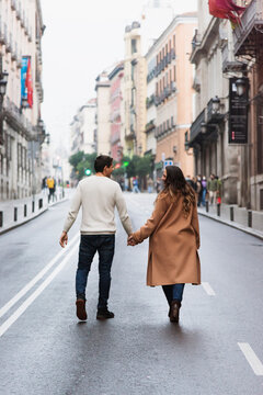 Full length content young couple in warm clothes holding hands and walking together on empty road in contemporary city on clear autumn day