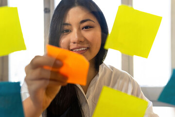 Young glad ethnic female entrepreneur arranging colorful paper stickers on transparent surface in office in daytime