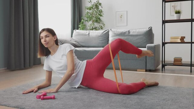 Young fitness woman lying on floor and training legs with resistance band. Attractive lady in activewear doing fitness exercises at home.