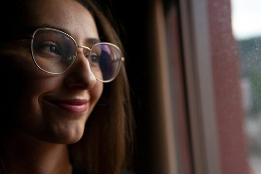 Crop positive young female with dark hair wearing eyeglasses standing in dark room and looking out window with smile
