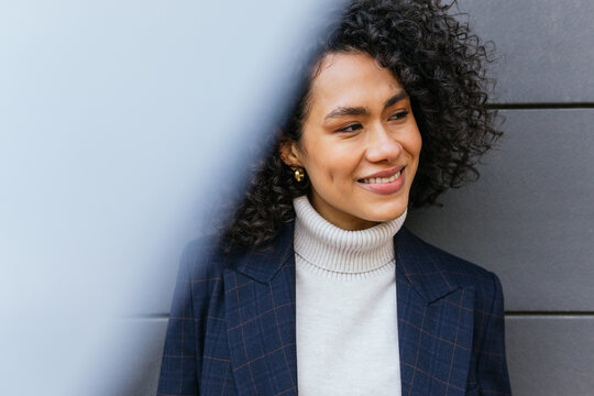 Delighted self assured young ethnic female in stylish clothes looking away while standing against gray striped wall