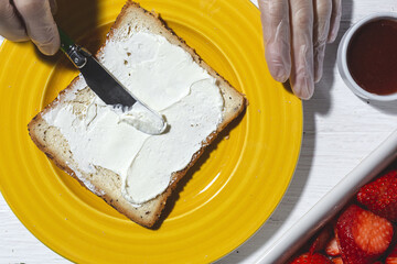 Top view crop anonymous female cook in latex gloves spreading yummy cream cheese on bread slice placed on yellow plate near cut strawberries