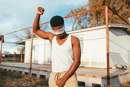 Anonymous masculine ethnic male protester in undershirt and blindfold standing with raised arm in countryside