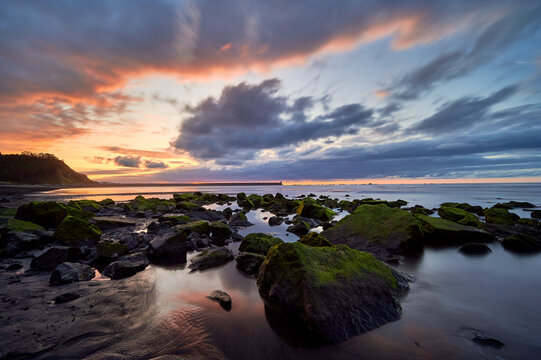 Picturesque view of boulders covered with moss on sandy beach of sea under cloudy sunset sky
