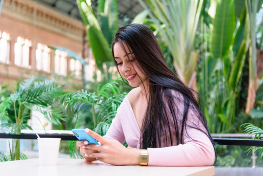 Confident young ethnic female with long dark hair in casual clothes smiling while sitting in cafe near green lush tropical plants and messaging on mobile phone