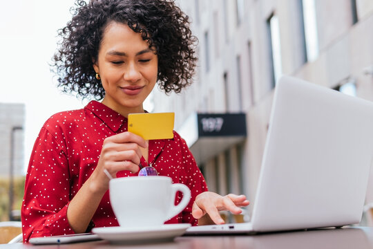 Happy young ethnic female with credit card in hand using laptop and doing online purchase on internet shop while sitting at table in outdoor cafe