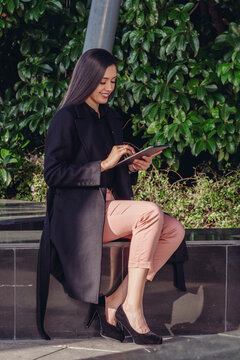 Full body of smiling young female in stylish outfit sitting on border on terrace and browsing tablet while checking information for business project