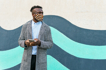 Anonymous contemplative ethnic male manager in ornamental mask with cellphone looking away near urban wall during COVID 19 pandemic